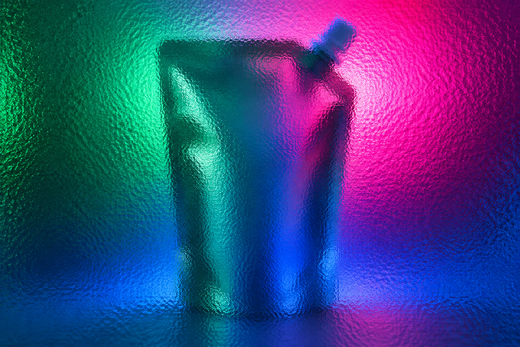 Unbranded spouted cosmetic refill pouch behind textured glass in vivid coloured light.