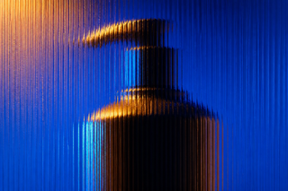 Unbranded pump-bottle prototype seen through full-frame vertical reeded glass in cobalt, cyan and amber light.