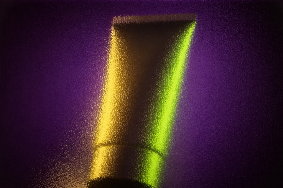 Unbranded squeeze tube seen through full-frame smoky rippled glass in aubergine, lime and gold light.