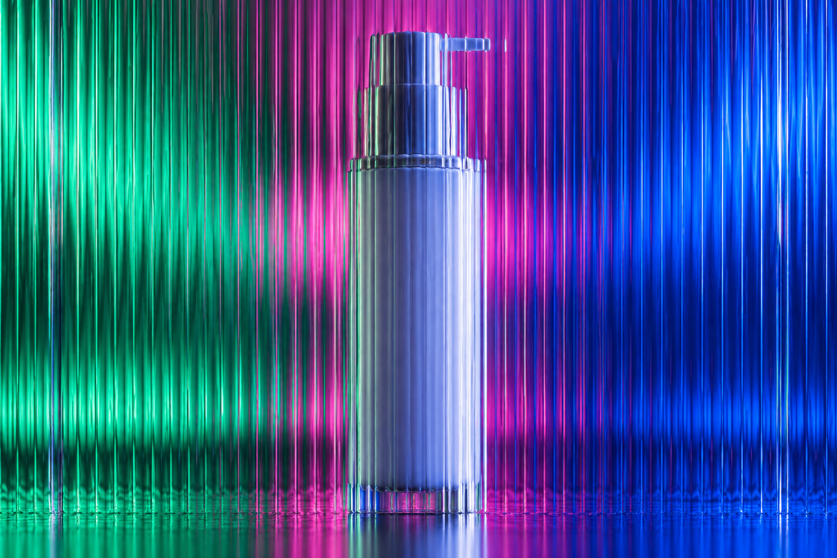 Unbranded airless pump bottle behind textured glass in vivid coloured light.