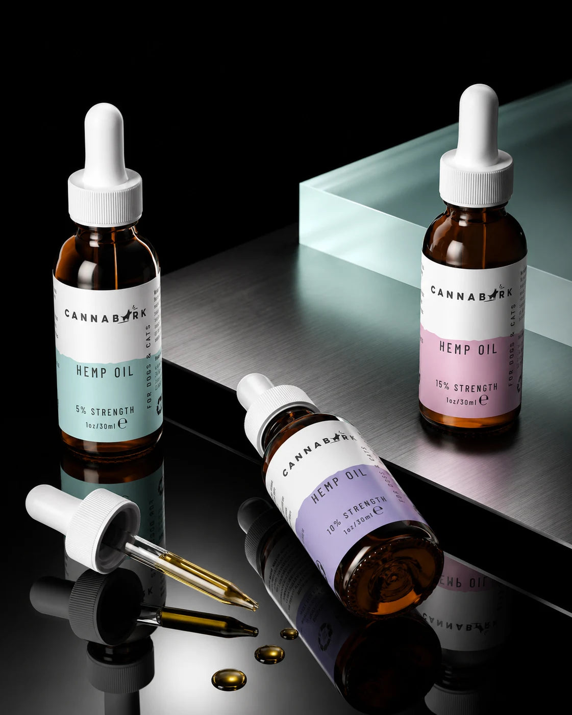 Three amber glass dropper bottles of Cannabior Hemp Oil with white caps are arranged on a reflective dark surface. One bottle is upright on the left with a pale blue label, one is upright on the right with a pale pink label, and one lies on its side in the foreground with a pale purple label.