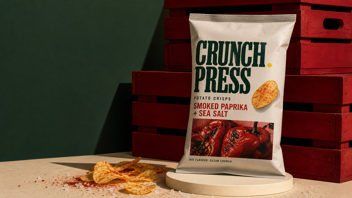Single Crunch Press potato crisp bag on a pedestal beside stacked red produce crates and scattered paprika crisps.